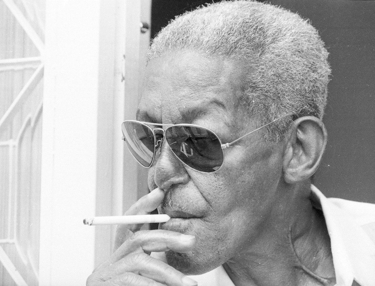 Homem negro de cabelos curtos e brancos, usando óculos de sol e fumando cigarro.
