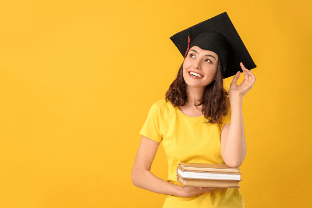 Estudante usando chapéu de graduação, segurando livros em sua mão, em fundo amarelo. 