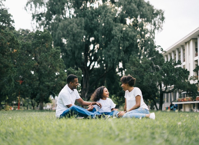 Um pai, uma mãe e a filha sentados sob a grama rindo e conversando
