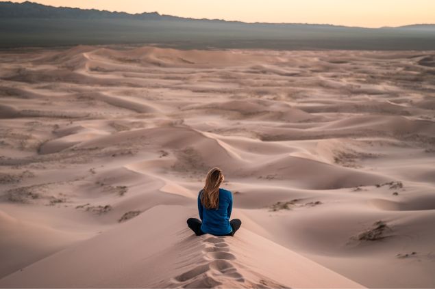 Mulher sentada na areia, pensando, em uma paisagem desértica.