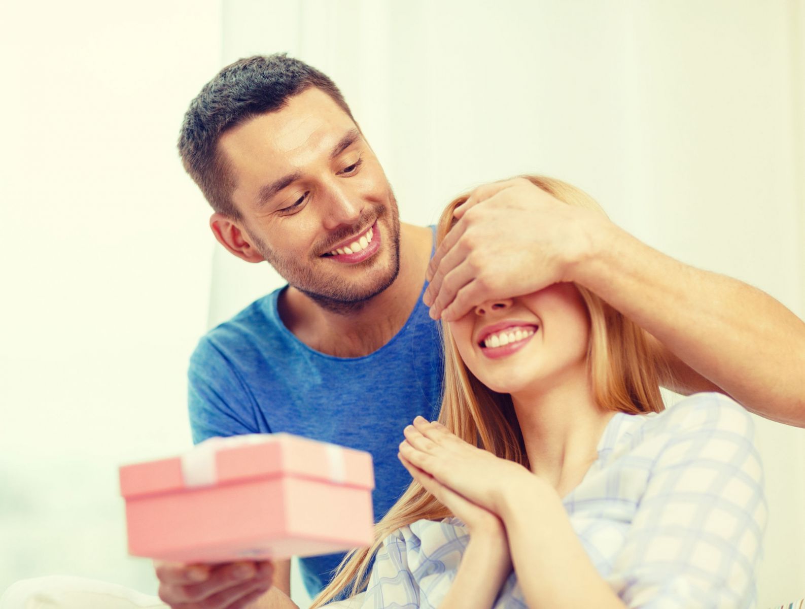 Homem cobrindo os olhos de uma mulher com uma mão, e segurando uma caixa de presente na frente dela com a outra mão.