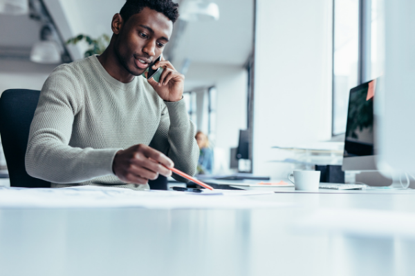 Homem falando ao telefone enquanto anota algo com a mão direita sob sua mesa de trabalho