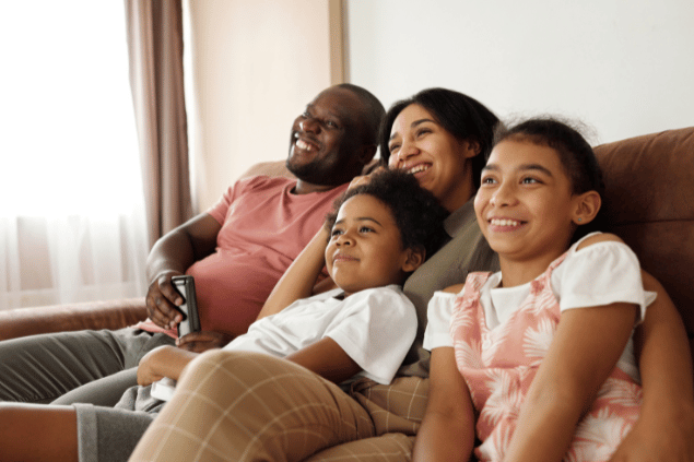 Uma família composta por um pai, uma mãe e duas meninas está sentada no sofá e sorrindo. Ao fundo, há uma sala.