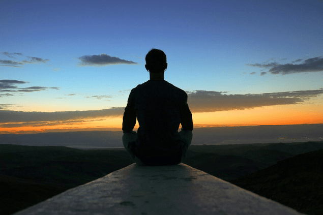 Um homem está sentado no concreto e olha para o horizonte. Ao fundo, há a natureza e um céu com um pôr do sol.