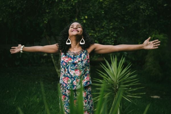 Mulher com os braços abertos, olhando pro alto e sorrindo no meio de plantas