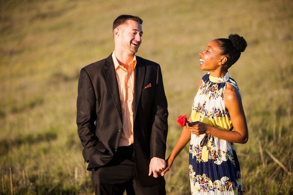 Casal de mãos dadas e sorrindo em um campo aberto. A mulher segura uma rosa vermelha