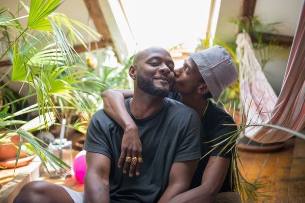 Casal abraçados em uma sala. Um deles está beijando a bochecha do outro