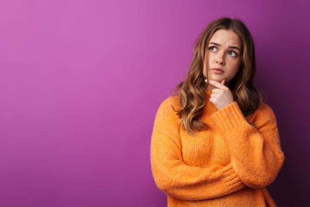 Imagem de uma moça de blusa laranja com a mão no queixo e semblante de dúvida em um fundo roxo.