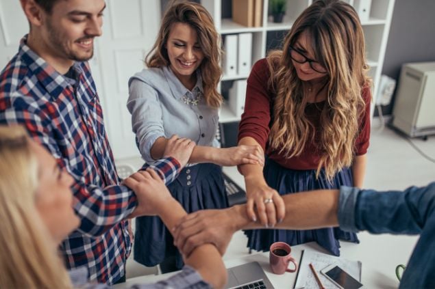 Imagem de um grupo de cinco pessoas que parecem fazer uma dinâmica corporal em um escritório