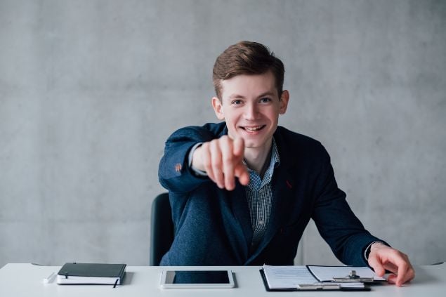 Imagem de um homem de terno sorrindo de frente para a tela, apontando para quem está olhando