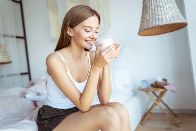 Imagem de uma mulher sentada na sala do sofá, com uma caneca de café, sorrindo de olhos fechados