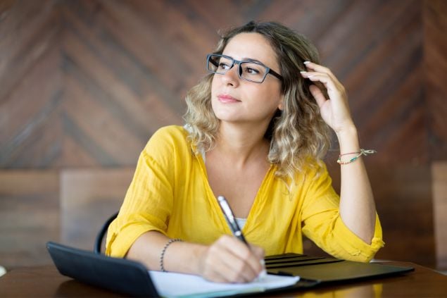 Imagem de uma mulher na frente do computador olhando para o lado, com uma caneta na mão e um caderno na mesa, com um sorriso no rosto como se estivesse tendo uma ideia