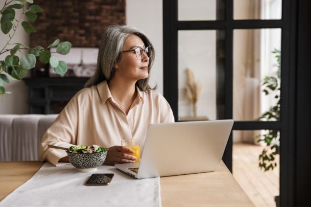 Mulher na frente de seu notebook com um copo de suco na mão e olhando para o lado