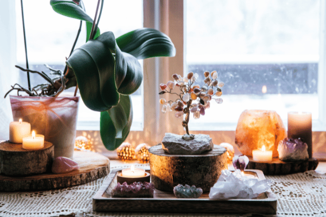 Um altar de Feng Shui composto por pedras, plantas e velas está no centro da imagem. Ao fundo, há uma janela.