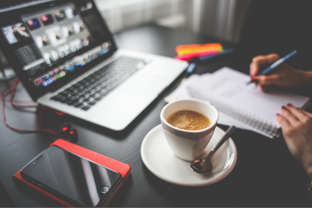 Uma pessoa escrevendo em um caderno em cima de uma mesa de trabalho com um computador, um celular e uma xícara de café