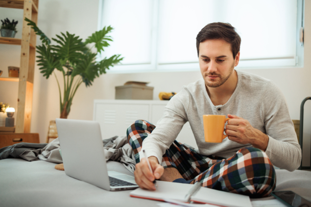 Homem sentado em cima da cama enquanto escreve em um caderno e toma um café