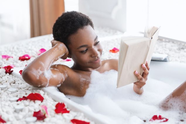 Imagem de uma mulher negra,  deitada em uma banheira de espuma, lendo um livro e fazendo um procedimento de spa, autocuidado e descanso.