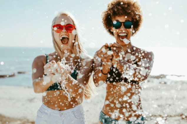 Imagem de duas mulheres na areia da praia brincado com bolhas de água. Elas estão sorrindo e felizes, diversão e integração da cunhada que é do signo de sagitário.