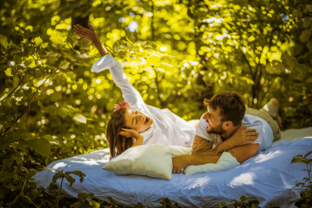 Imagem de um casal deitado sobre um colchão em meio a natureza, trazendo o conceito de fazer algo diferente para o relacionamento não cair na rotina.