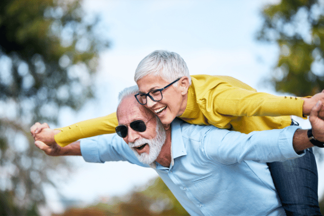 Imagem de um casal 50+ felizes e sorridentes em uma praça arborizada.