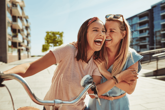 Imagem de duas mulheres caminhando. Uma delas empurra sua bicicleta. Elas estão sorrindo e felizes, simbolizando o respeito que elas nutrem uma pela outra.