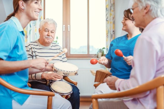 Imagem de um casal 60+ em um sala de casa praticando a musicoterapia com assistentes de saúde.