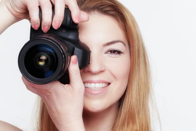 Imagem de uma mulher loura de cabelos longos, segurando uma câmera fotográfica com as suas mãos.