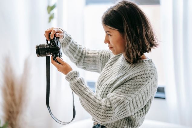 Imagem de uma mulher fotógrafa usando um suéter de linha cinza claro, segurando um câmera fotográfica em suas mãos.