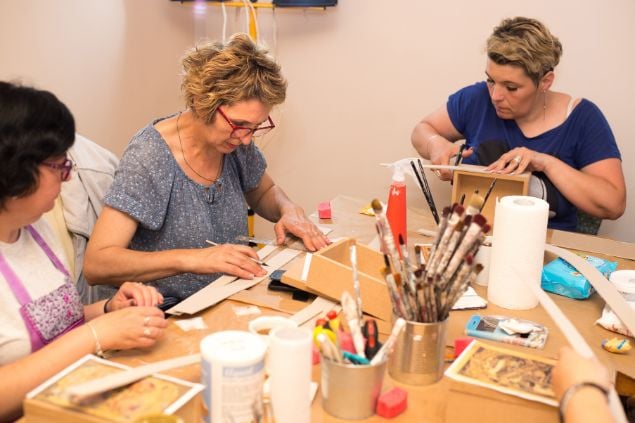 Imagem de três mulheres sentadas fazendo aula de artes. Sobre uma mesa de madeira, vários itens como lápis, canetas, pincéis, complementam a foto.