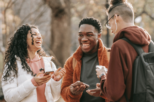 Um grupo de três amigos interage e sorri. Eles estão com seus celulares na mão. Ao fundo, há um parque desfocado.