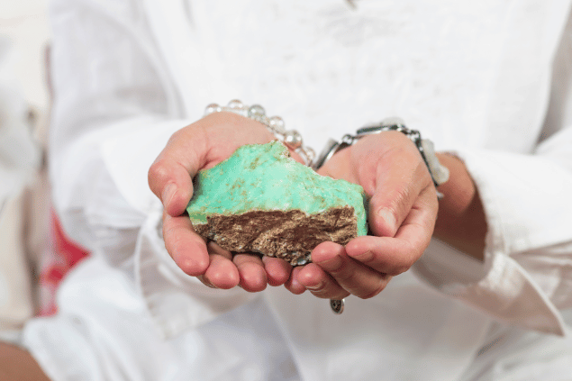 As duas mãos de uma mulher seguram uma pedra verde. Ela veste uma blusa branca e usa pulseiras de pedras.