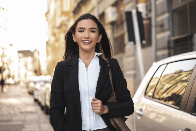 Uma mulher de cabelos castanhos que veste roupas formais está sorrindo. A imagem sinaliza que a mulher está no mercado de trabalho e é independente. Ao fundo, há uma cidade desfocada.