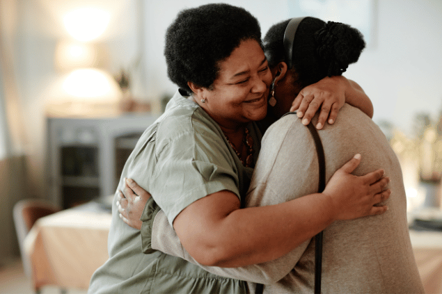 Duas mulheres negras se abraçam, sinalizando uma ideia de apoio. Ao fundo, há uma sala desfocada.