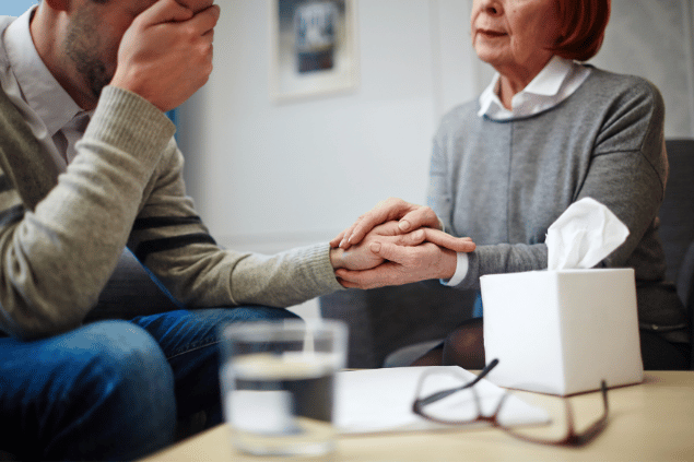 Uma senhora coloca as suas mãos sobre a mão de um homem que aparenta não estar bem emocionalmente. Ela oferece apoio.