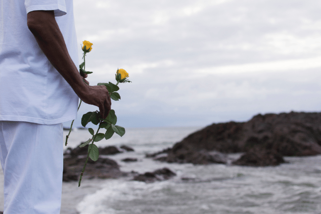 Um participante da religião candomblé está de frente para o mar e segura duas flores amarelas que irá ofertar no local. Ele usa roupas brancas.