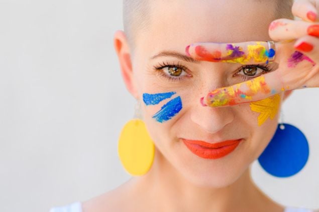 Mulher fazendo sinal de V nos olhos, com a mão e o rosto manchados de tintas coloridas.