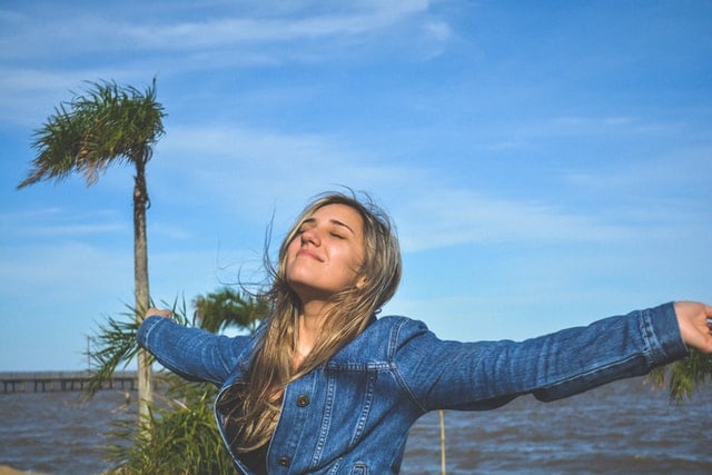 Mulher branca de braços abertos próxima à praia.