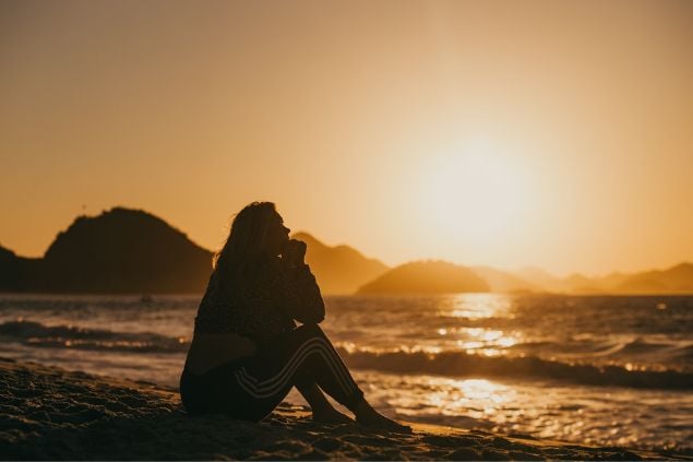 Mulher sentada na praia, observando o mar no pôr do Sol.