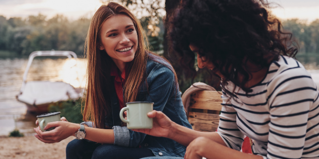 Duas mulheres sorriem enquanto conversam em ambiente externo. Elas seguram canecas. 