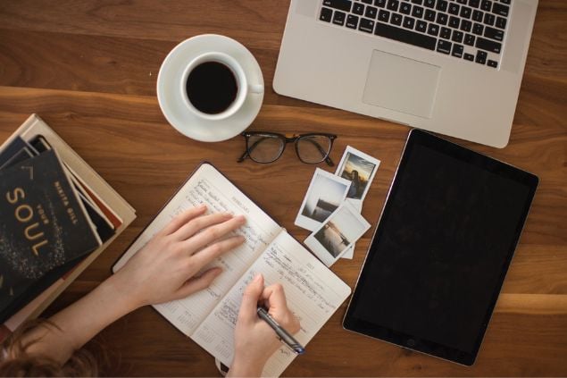 Mão feminina sobre uma agenda aberta. Ao redor, livros, um óculos, uma xícara de café, um notebook, um tablet e fotografias.