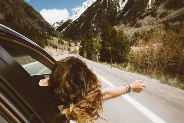 Mulher com a cabeça para fora da janela do carro e o braço esticado, aproveitando uma viagem pela estrada.