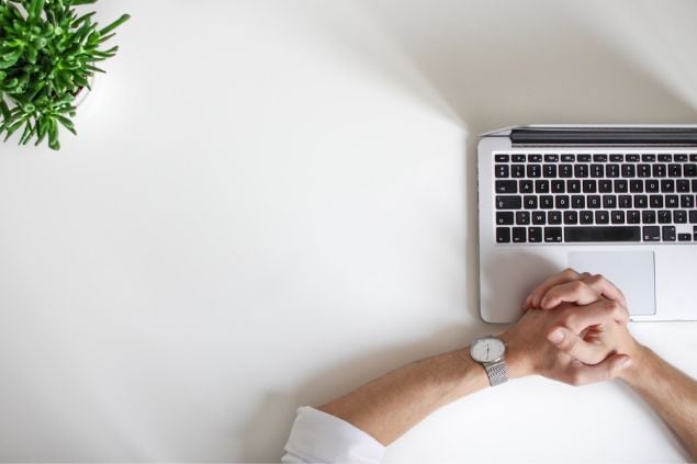 Mãos masculinas cruzadas sobre uma mesa branca, à frente de um notebook aberto. Uma plantinha aparece parcialmente no canto superior esquerdo da imagem.